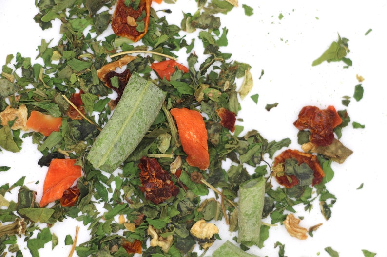May include: A close-up of a mix of dried herbs and vegetables, including green leafy herbs, orange and red dried fruit pieces, and some larger green pieces.