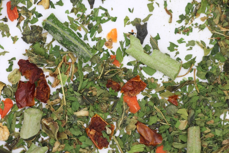 May include: A close-up of a mix of dried herbs and vegetables, including green leafy herbs, dried tomatoes, and orange carrot pieces.