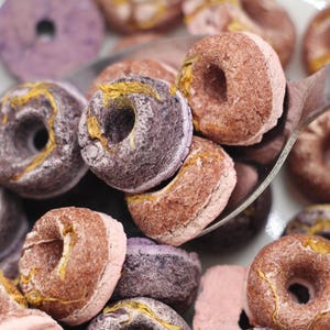 K&ouml;nnte beinhalten: Eine Sch&uuml;ssel mit lila und braunen donutf&ouml;rmigen Cerealien. Das M&uuml;sli befindet sich in einer wei&szlig;en Sch&uuml;ssel mit einem silbernen L&ouml;ffel. Das M&uuml;sli ist ein Fr&uuml;hst&uuml;cksessen.
