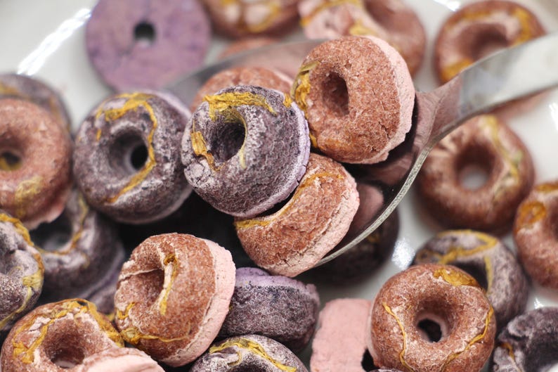 K&ouml;nnte beinhalten: Eine Sch&uuml;ssel mit lila und braunen donutf&ouml;rmigen Cerealien. Das M&uuml;sli befindet sich in einer wei&szlig;en Sch&uuml;ssel mit einem silbernen L&ouml;ffel. Das M&uuml;sli ist ein Fr&uuml;hst&uuml;cksessen.