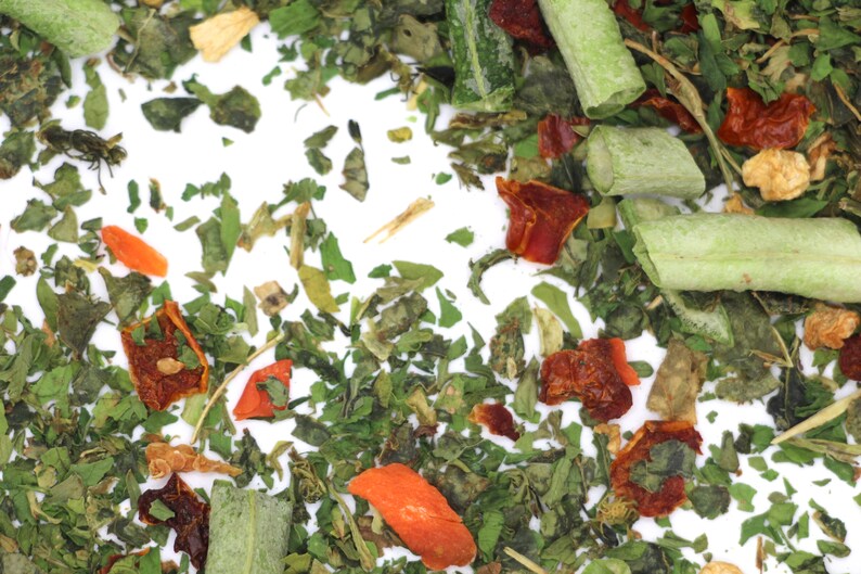 May include: A close-up of a mix of dried herbs and vegetables, including green leafy herbs, red and orange dried peppers, and long green pieces of dried vegetables.
