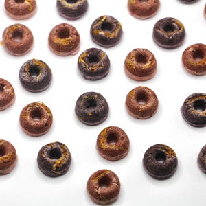 K&ouml;nnte beinhalten: Eine Sammlung kleiner, runder, donutf&ouml;rmiger Cerealienst&uuml;cke in verschiedenen Farben, darunter Braun, Schwarz und Gold. Die Cerealienst&uuml;cke sind auf einer wei&szlig;en Oberfl&auml;che angeordnet.