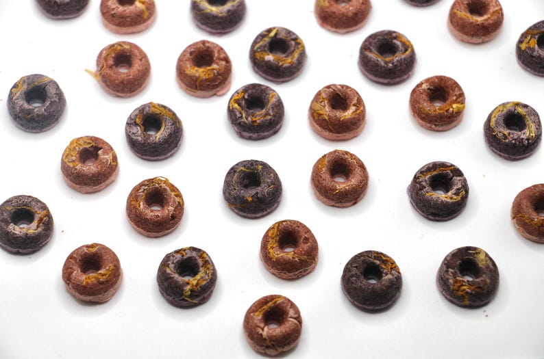 K&ouml;nnte beinhalten: Eine Sammlung kleiner, runder, donutf&ouml;rmiger Cerealienst&uuml;cke in verschiedenen Farben, darunter Braun, Schwarz und Gold. Die Cerealienst&uuml;cke sind auf einer wei&szlig;en Oberfl&auml;che angeordnet.