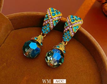 May include: A pair of gold-toned drop earrings with teal and purple accents. The earrings feature a geometric design with teal-colored stones and a large, faceted blue crystal drop. The text "WM ACC" and "LUXURY FASHION" is visible.