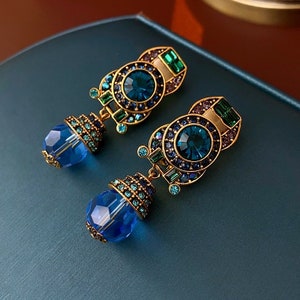 Puede incluir: Un par de pendientes colgantes de tono dorado con detalles de cristal azul y verde. Los pendientes presentan un diseño redondo y geométrico con un gran cristal azul en el centro.