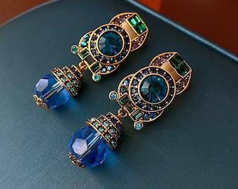 May include: A pair of gold tone dangle earrings with blue and green crystal accents. The earrings feature a round, geometric design with a large blue crystal in the center.