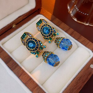 Puede incluir: Un par de pendientes dorados adornados con piedras preciosas azules y verdes. Cada pendiente presenta una gran piedra azul redonda rodeada de pequeños detalles azules y verdes, y un cristal azul facetado colgante. Los pendientes se muestran en una caja de joyería de madera.
