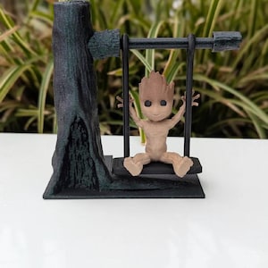 May include: A 3D-printed figurine of a character from Guardians of the Galaxy, sitting on a swing. The swing is attached to a tree trunk. The figurine is light brown, and the tree and swing are dark green and black.