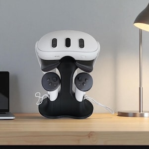 May include: A white and black VR headset and controllers are stored in a black stand. The VR headset has three black sensors on the front. The controllers are white with black buttons. The stand is on a wooden surface.