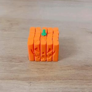 May include: A cube-shaped Halloween decoration in the shape of a carved pumpkin. The pumpkin is orange with a green stem and carved facial features. The decoration is made of vertical orange blocks.