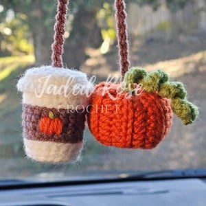 May include: Crocheted car charm featuring a coffee cup and a pumpkin. The coffee cup is white, brown, and orange, with a small pumpkin applique. The pumpkin is orange with green leaves. The charm hangs from a brown string.