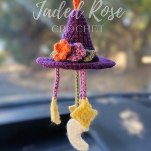 Op de afbeelding: Een paarse gehaakte heksenhoed auto-hanger met oranje en paarse bloemen, gele sterren en een witte maan. De hanger wordt opgehangen met een oranje koord en heeft de tekst "Jaded Rose Crochet".