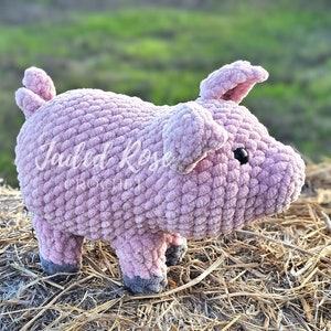 May include: A pink crocheted pig toy with black eyes. The pig is standing on a bed of straw.
