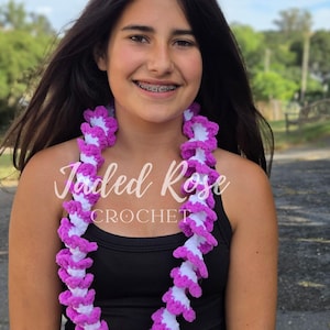 May include: A handmade crochet lei featuring alternating sections of white and vibrant purple. The lei is displayed against a natural outdoor backdrop. The text "Jaded Rose CROCHET" is visible on the image.