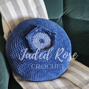 Puede incluir: Una almohada de arándano azul tejida a crochet con una flor blanca y azul tejida a crochet en el centro. La almohada está sentada sobre una manta a rayas gris y blanca.