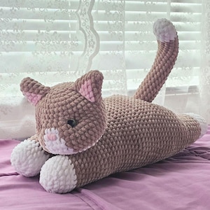 May include: A handmade crochet cat plushie in shades of brown, pink, and white. The cat is lying down with its tail curved upwards. The plushie has a textured appearance and is resting on a purple surface.