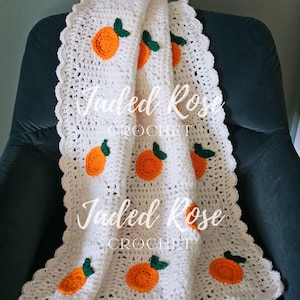 Puede incluir: Una manta blanca de ganchillo con frutas naranjas y detalles de hojas verdes. La manta tiene un borde festoneado y está sobre una silla verde oscuro. El texto "Jaded Rose Crochet" es visible.