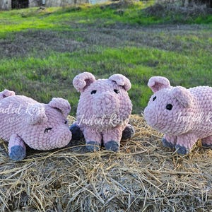 May include: Three crocheted pig plushies in a pink color with black eyes. The plushies are sitting on a bed of hay.