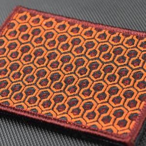 Peut inclure: Un patch tissé rouge, orange et noir avec un motif géométrique répétitif d'hexagones. Le patch a une bordure bordeaux.