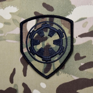 Puede incluir: Un parche bordado en blanco y negro con el símbolo imperial de Star Wars. El parche tiene forma de escudo y está sobre un fondo de camuflaje.