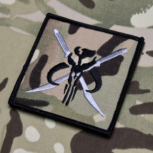 May include: A black and white embroidered patch with a Mandalorian symbol and crossed lightsabers on a camouflage background.