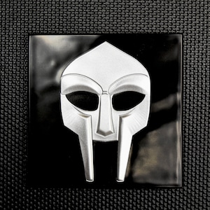May include: A silver metal mask with a distinctive design, featuring a pointed top, eye holes, and a prominent nose guard. The mask is set against a black background.