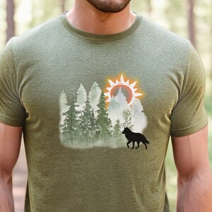 May include: A green t-shirt with a graphic print of a wolf standing in a forest with a sun rising behind the trees.