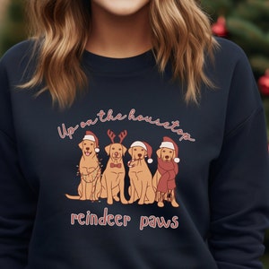 May include: Navy blue sweatshirt featuring a festive design. The graphic shows four golden retriever dogs dressed in holiday attire, including Santa hats and reindeer antlers. The text "Up on the housetop reindeer paws" is written above and below the dogs in a script font.