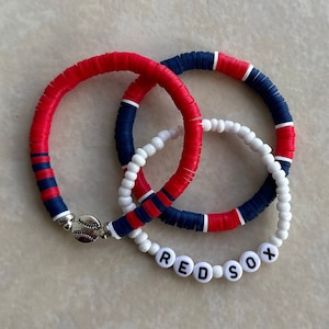 May include: Three bracelets, one red and white, one blue and white, and one white with the words "RED SOX" in black. The bracelets are made of beads and are stacked on top of each other.