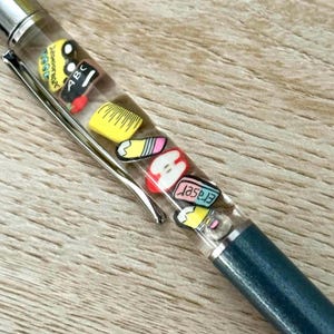 May include: A clear pen with a silver clip and a dark teal barrel. The pen's clear chamber is filled with miniature school-themed charms, including an apple, pencil, ruler, and eraser.