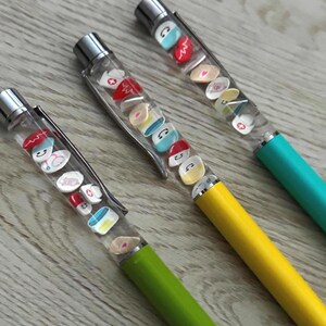 Op de afbeelding: Drie pennen met doorzichtige vaten gevuld met miniatuur medische bedels. De pennen hebben zilveren accenten en de vaten zijn gevuld met kleine pillen, stethoscopen en verpleegstershoeden. De penvaten hebben groene, gele en blauwgroene bases.