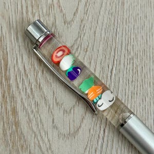 May include: Silver pen with a clear barrel filled with colorful miniature food-shaped charms. The charms include a tomato slice, white egg, purple eggplant, green leaf, and sushi-like shapes. The pen has a silver clip and tip.