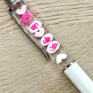 May include: A white pen with a clear barrel filled with pink heart and elephant-shaped charms. The pen has a silver clip and tip, and is resting on a light brown wooden surface.