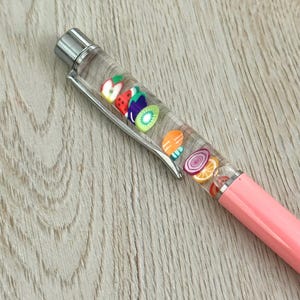 May include: A pink pen with a clear barrel filled with colorful fruit-shaped charms, including strawberries, kiwi, and oranges. The pen has a silver clip and tip, and is resting on a light wood surface.
