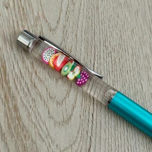 May include: A teal pen with a clear barrel filled with colorful fruit-shaped charms. The pen has a silver clip and tip, and the fruit charms include strawberries, oranges, kiwi, and grapes. The pen rests on a light wood-grain surface.
