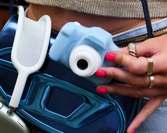 Könnte beinhalten: Eine blaue Spielzeugkamera mit einem weißen Objektiv wird in der Hand einer Person gehalten. Die Kamera ist an einer blauen metallischen Tasche befestigt, an der ein weißer Netzbeutel hängt.