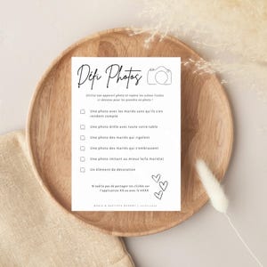 May include: A white card with black text on a wooden plate. The card has a title 'Defi Photos' and a list of photo challenges for a wedding. The challenges include: 'Une photo avec les mariés sans qu'ils s'en rendent compte', 'Une photo drôle avec toute votre table', 'Une photo des mariés qui rigolent', 'Une photo des mariés qui s'embrassent', 'Une photo imitant au mieux le/la marié(e)', and 'Un élément de décoration'. The card also includes the text 'N'oublie pas de partager tes clichés sur l'application XX ou avec le #XXX' and 'MARIS & SARTISTE DUPONT | 01/01/2023'.
