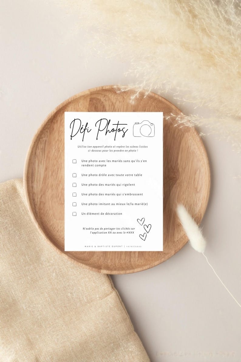 May include: A white card with black text on a wooden plate. The card has a title 'Defi Photos' and a list of photo challenges for a wedding. The challenges include: 'Une photo avec les mariés sans qu'ils s'en rendent compte', 'Une photo drôle avec toute votre table', 'Une photo des mariés qui rigolent', 'Une photo des mariés qui s'embrassent', 'Une photo imitant au mieux le/la marié(e)', and 'Un élément de décoration'. The card also includes the text 'N'oublie pas de partager tes clichés sur l'application XX ou avec le #XXX' and 'MARIS & SARTISTE DUPONT | 01/01/2023'.