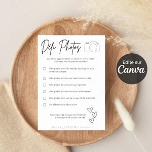 May include: A white card with black text and a simple drawing of a camera. The card is titled 'Difi Photos' and has a list of photo challenges for a wedding. The text on the card reads: 'Utilise ton appareil photo et repère les scènes listées ci-dessous pour les prendre en photo!'  'Une photo avec les mariés sans qu'ils s'en rendent compte'  'Une photo drôle avec toute votre table'  'Une photo des mariés qui rigolent'  'Une photo des mariés qui s'embrassent'  'Une photo imitant au mieux le/la marié(e)'  'Un élément de décoration'  'N'oublie pas de partager tes clichés sur l'application XX ou avec le #XXX'  'MARIE & BAPTISTE DURORT | 16/07/2022'  The card also has the text 'Edite sur Canva' in a black circle.