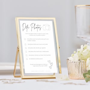 May include: A gold-framed photo stand with a white card inside. The card has black text that reads 'Defi Photos' and includes a list of photo challenges for wedding guests. The text on the card reads: 'Utilise ton appareil photo et repère les scènes listées ci-dessous pour les prendre en photo!' followed by a list of photo challenges: 'Une photo avec les mariés sans qu'ils s'en rendent compte', 'Une photo drôle avec toute votre table', 'Une photo des mariés qui rigolent', 'Une photo des mariés qui s'embrassent', 'Une photo imitant au mieux le/la marié(e)', 'Un élément de décoration'. The card also includes a reminder to share photos on the app XXX and the names of the bride and groom, Marie & Baptiste Dupont.