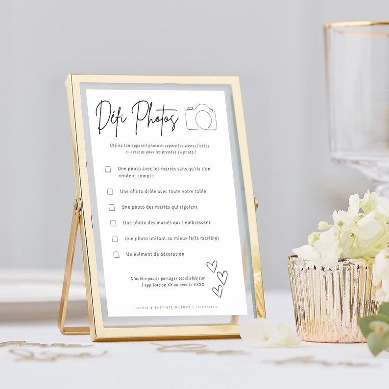 May include: A gold-framed photo stand with a white card inside. The card has black text that reads 'Defi Photos' and includes a list of photo challenges for wedding guests. The text on the card reads: 'Utilise ton appareil photo et repère les scènes listées ci-dessous pour les prendre en photo!' followed by a list of photo challenges: 'Une photo avec les mariés sans qu'ils s'en rendent compte', 'Une photo drôle avec toute votre table', 'Une photo des mariés qui rigolent', 'Une photo des mariés qui s'embrassent', 'Une photo imitant au mieux le/la marié(e)', 'Un élément de décoration'. The card also includes a reminder to share photos on the app XXX and the names of the bride and groom, Marie & Baptiste Dupont.