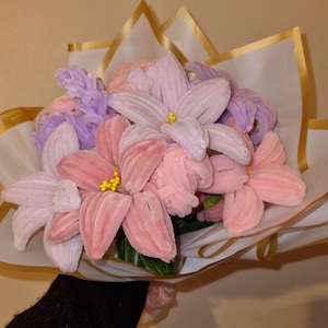 Op de afbeelding: Een boeket handgemaakte bloemen in roze en paarse tinten. De bloemen zijn verpakt in wit papier met gouden rand. De bloemblaadjes hebben een zachte, fluweelzachte textuur en de centra hebben gele accenten.