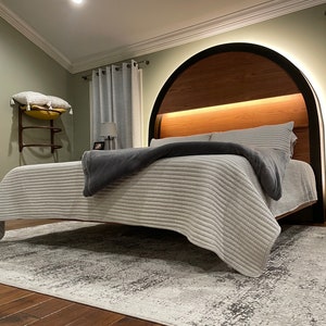 May include: A bedroom with a modern bed featuring a curved wooden headboard with integrated lighting. The bed has a white and gray striped duvet and a gray throw blanket. The room has a light green wall, a rug, and a wooden nightstand.