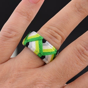 May include: A green, white, and black finger ring made of braided fabric. The ring is worn on a person's finger.