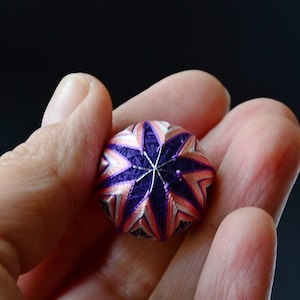 Op de afbeelding: Een klein, rond, stervormig, handgestikt ornament in paars en roze.