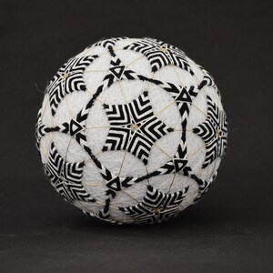 Peut inclure: Une boule Temari sphérique, faite à la main. Elle présente un motif géométrique complexe en noir et blanc, avec des formes en étoile. Des fils dorés accentuent le design, créant une pièce détaillée et visuellement frappante.