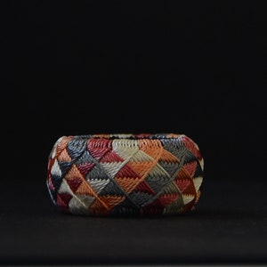 May include: A colorful, woven ring with a geometric pattern in shades of red, orange, gray, and white. The ring is made of a tightly woven material and has a smooth, rounded surface.