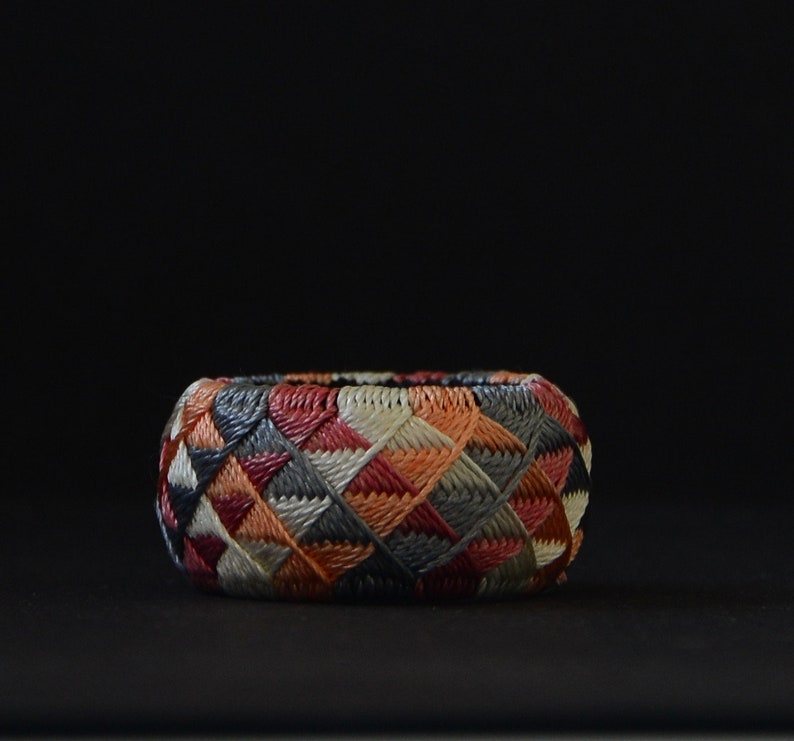 May include: A colorful, woven ring with a geometric pattern in shades of red, orange, gray, and white. The ring is made of a tightly woven material and has a smooth, rounded surface.