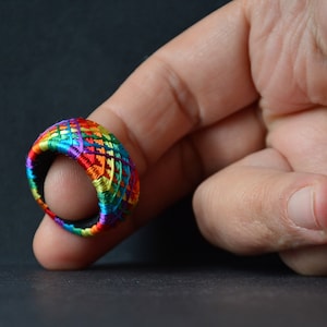 Könnte beinhalten: Ein bunter Regenbogen-Fingerhut aus gewebtem Garn. Der Fingerhut wird an einem Finger getragen.