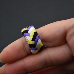 May include: A purple, yellow, white and black braided ring worn on a finger.